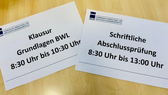Zwei Beschilderungen auf weißen A4-Blättern: Die erste kündigt eine Klausur in Grundlagen BWL an, die von 8:30 bis 10:30 Uhr stattfindet. Die zweite informiert über eine schriftliche Abschlussprüfung, die von 8:30 bis 13:00 Uhr dauert.