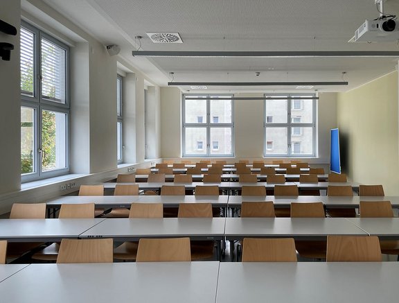 Ein heller Klassenraum mit vielen langen Tischen und Stühlen. Die Tische sind in mehreren Reihen angeordnet, und große Fenster lassen Tageslicht herein. An einer Wand befindet sich ein Bildschirm. Der Raum wirkt ordentlich und bereit für Unterricht oder Seminare.