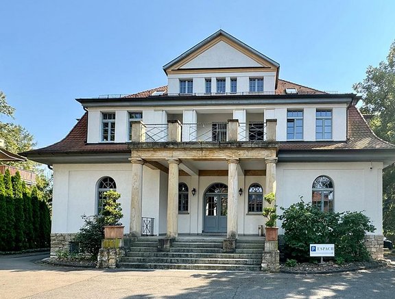 Eine elegante Villa mit weißen Wänden und einem roten Ziegeldach. Die Front wird von Säulen getragen, und es gibt große Fenster. Treppen führen zur Eingangstür. Vor der Villa befinden sich bepflanzte Blumenkübel und ein Schild mit dem Schriftzug „ESPACH“.