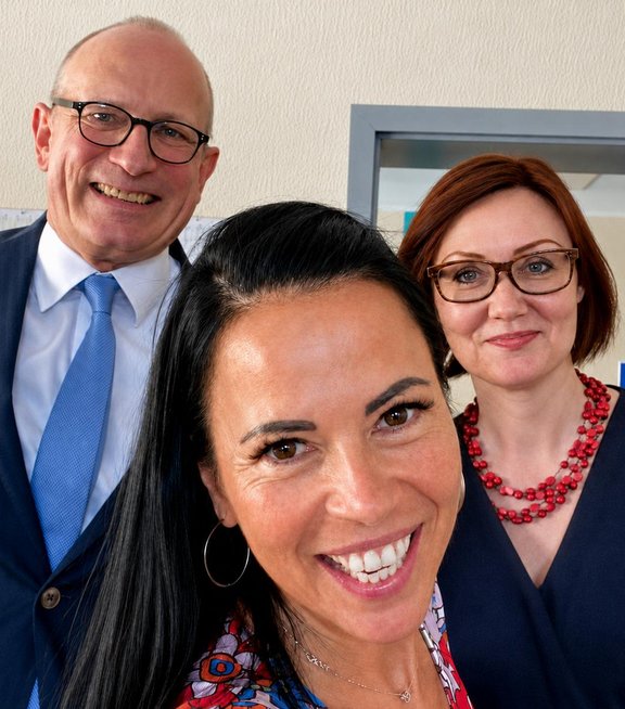 Drei Personen stehen in einem Raum und schauen in die Kamera. Eine Frau mit langen, dunklen Haaren und einem bunten Oberteil lächelt. Hinter ihr stehen zwei Personen, die Leiterin der Geschäftsstelle Frau Susanne Pfeiffer mit einer mit Brille und in einem blauen Oberteil, die andere Person mit einem Hemd und Krawatte ist der Studienleiter Prof. Fischer, die Personen lächeln ebenfalls.