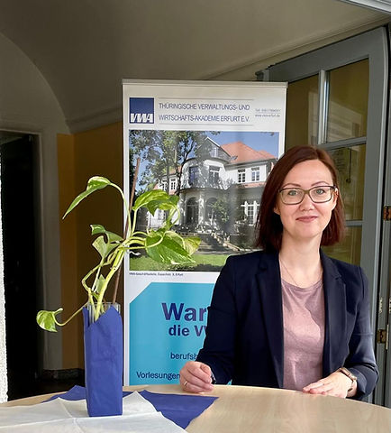 Eine Frau mit Brille sitzt an einem Tisch und lächelt. Vor ihr steht ein kleiner Pflanzenkübel. Im Hintergrund ist ein Banner der Thüringischen Verwaltungs- und Wirtschaftsakademie mit einem Gebäude zu sehen. Der Text auf dem Banner enthält Informationen über berufliche Vorlesungen.